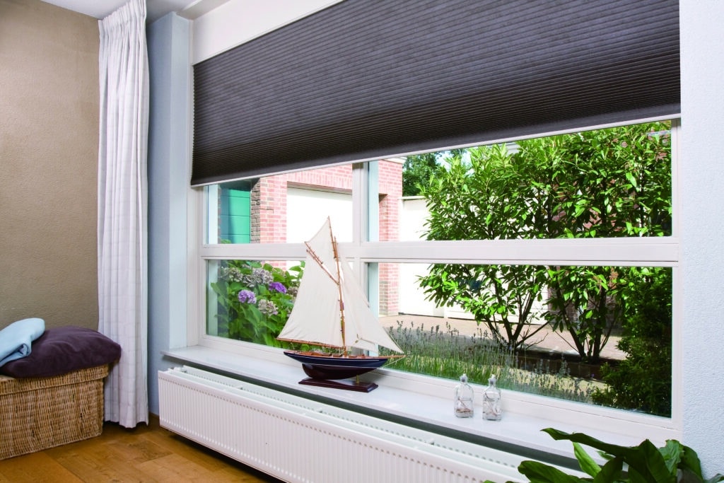 Living room window with dark honeycomb blinds in an Adelaide home, decorated with a model sailboat and garden view.