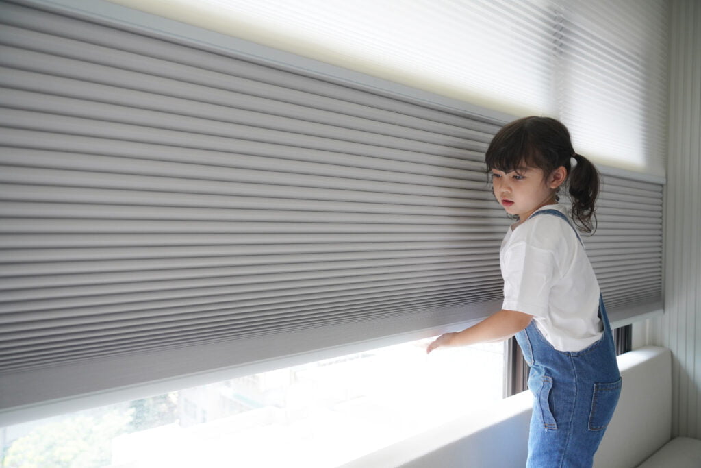 A young child standing near a window fitted with cordless grey Norman Honeycomb Blinds, highlighting a safe and modern window covering option.