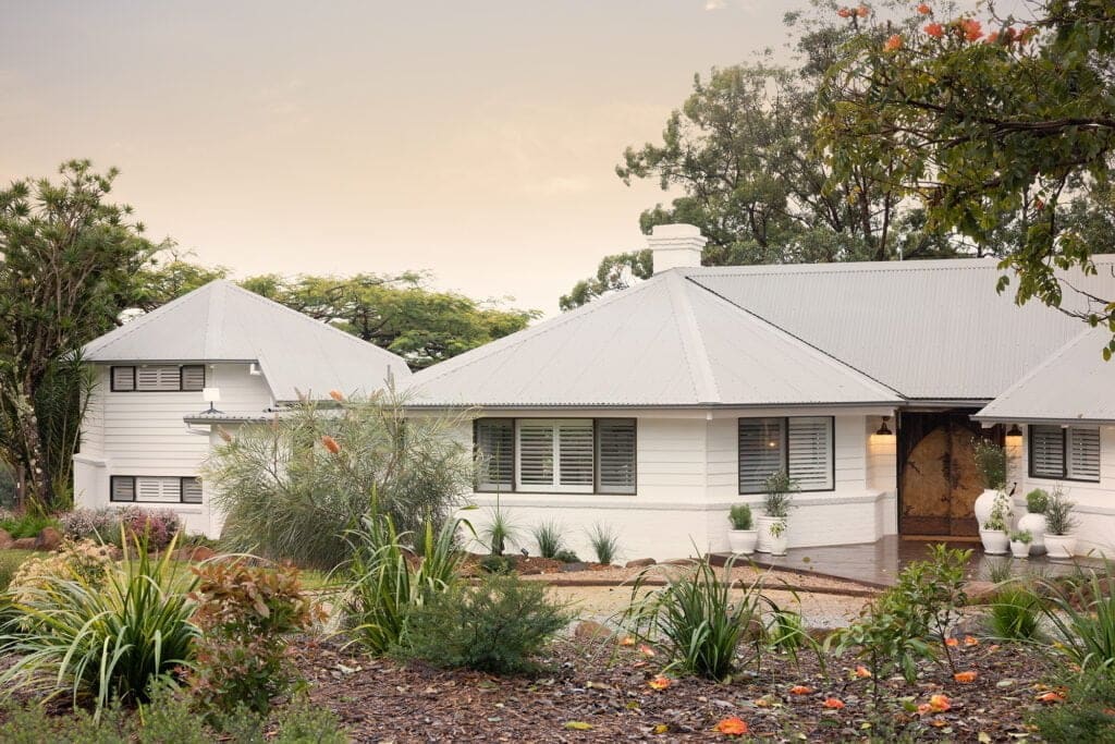 A charming white weatherboard home with grey roof and windows fitted with sleek white Norman Shutters, surrounded by lush native landscaping.