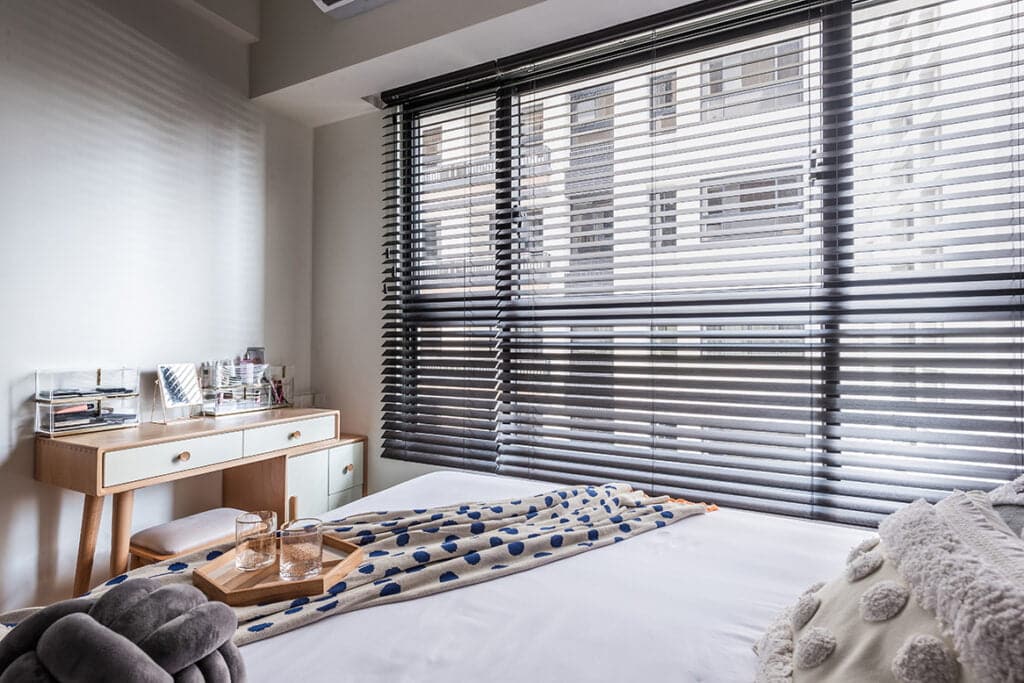 A bright bedroom interior featuring large windows fitted with sleek black Norman Venetian Blinds, a wooden vanity desk, and a neatly made bed with decorative throws and cushions.