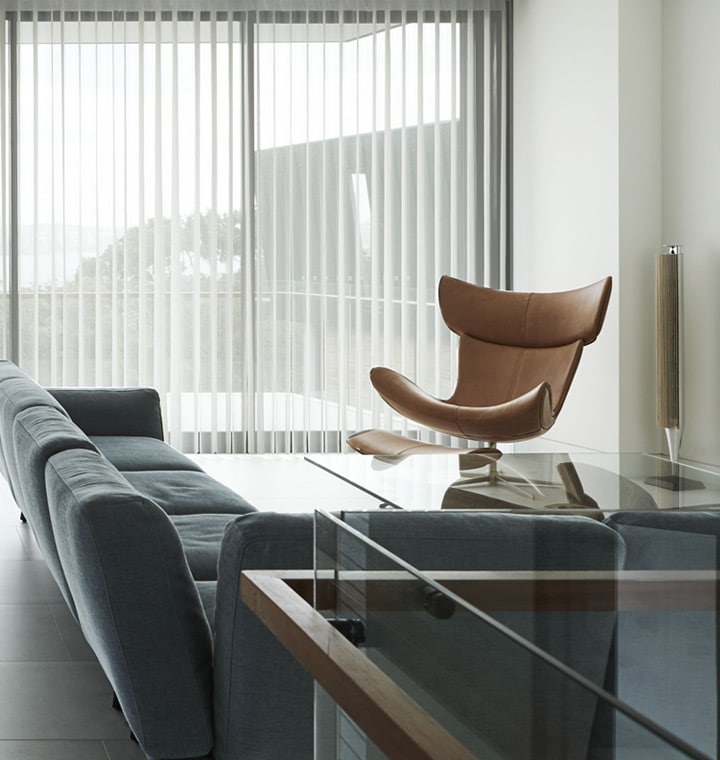 Modern living room with Veri Shades filtering light behind a leather lounge chair and grey sofa, vertical fabric panels providing soft daylight and privacy.