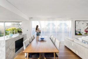 Woman adjusting Veri Shades in a sunlit open-plan dining and living room, sheer fabric panels filtering light for privacy and ambience.